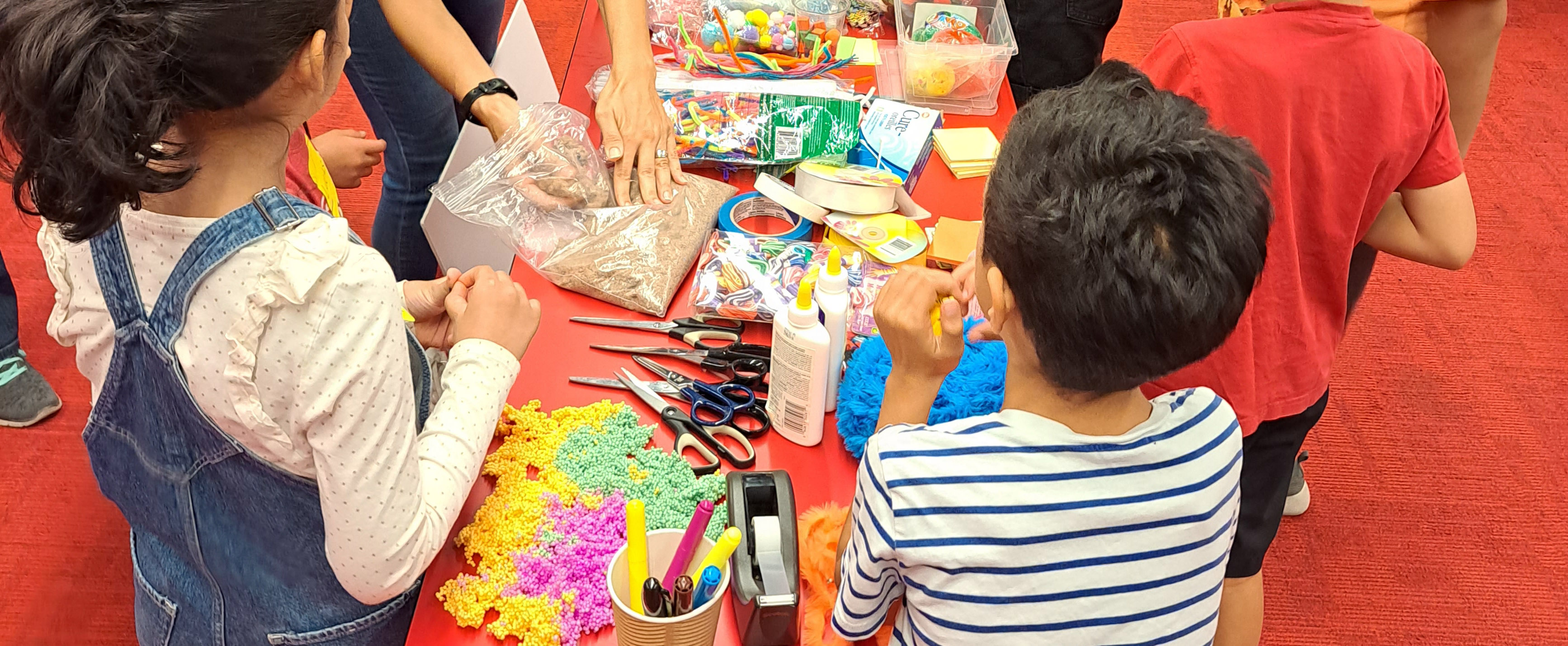 Art and craft tools and material on a red desk with kids standing around the table. 