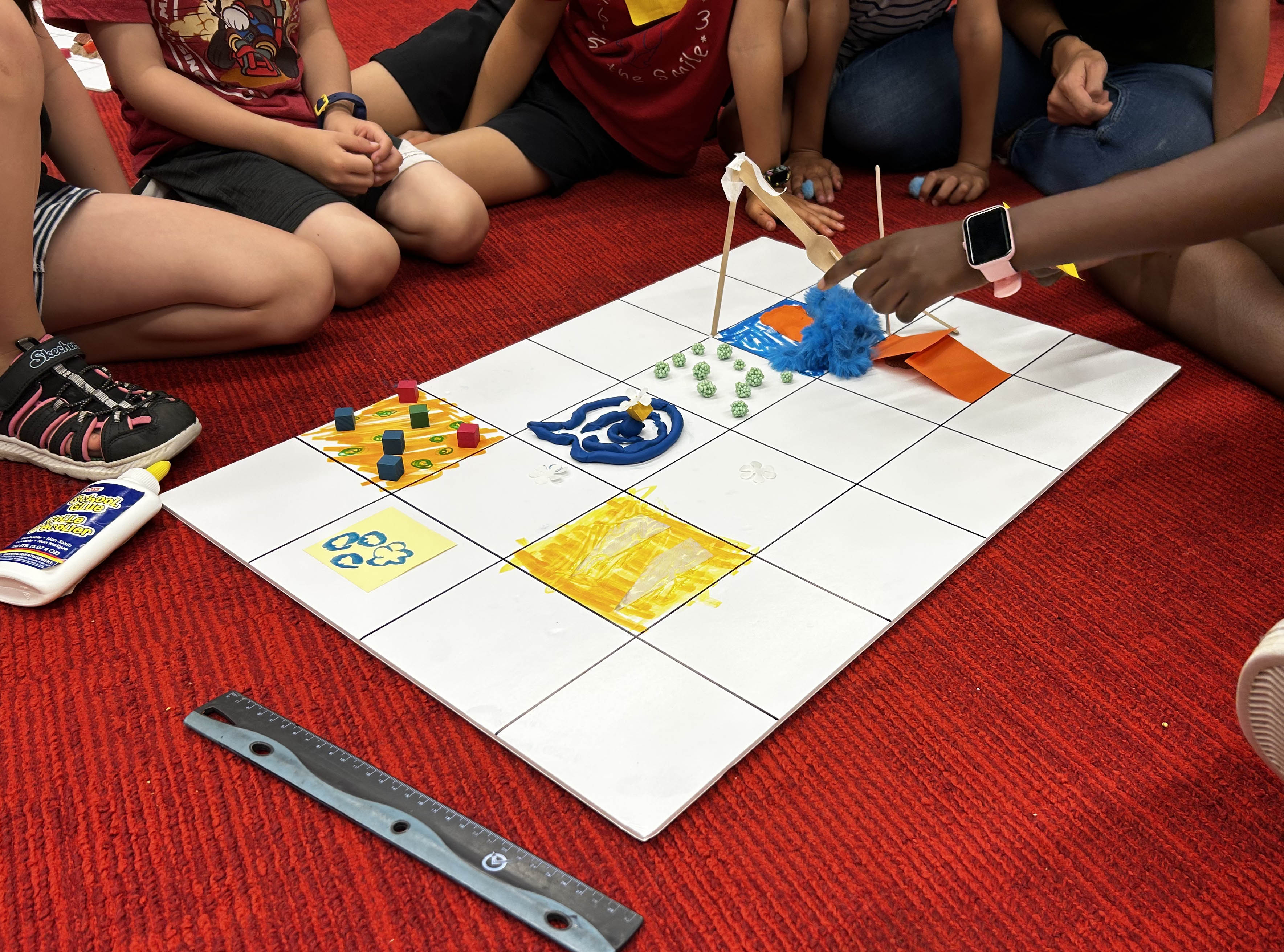 Kids sitting on the floor around a board and trying to design a customized grid on this board with paint and other craft materials. 