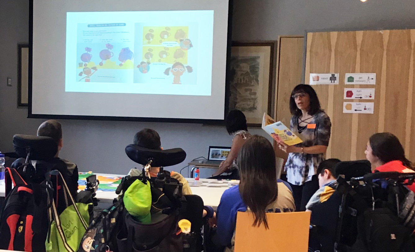 Woman reading a book for a group of children on mobility devices and projecting the page on the screen. 